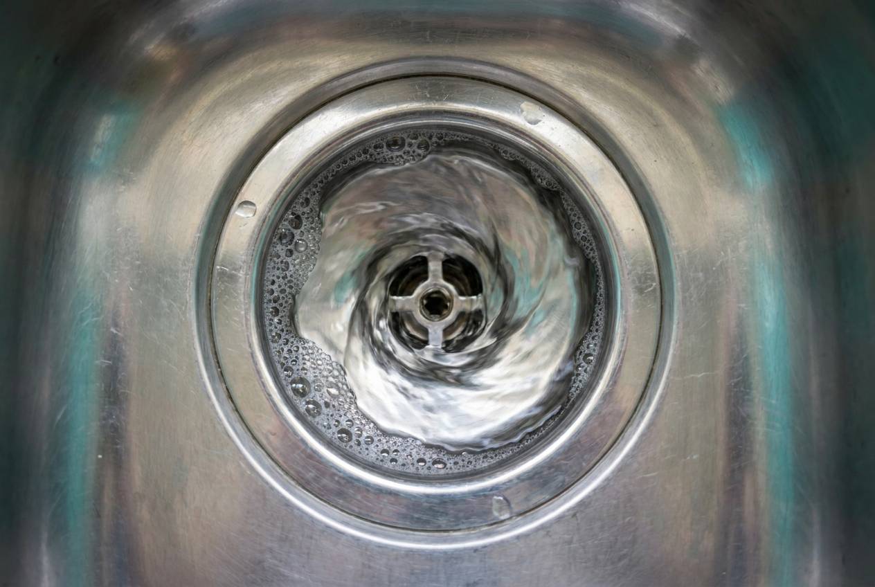 Immagine illustrativa: acqua che scende con un piccolo vortice regolare nello scarico di un lavello da cucina in acciaio.